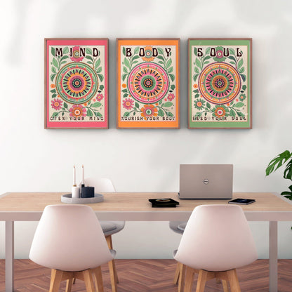 May include: Three framed prints with colorful floral designs and text. The first print has a pink background and the text &#39;MIND OPEN YOUR MIND&#39;. The second print has an orange background and the text &#39;BODY NOURISH YOUR BODY&#39;. The third print has a green background and the text &#39;SOUL FEED YOUR SOUL&#39;. The prints are hanging on a white wall above a wooden table with two white chairs.